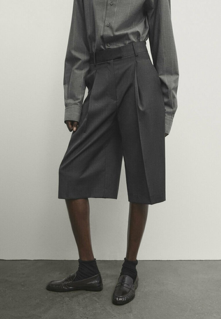 Culottes plissés gris avec une taille élégante, fabriqués à partir d'un tissu texturé. Porté avec des chaussures et des chaussettes noires, sur un fond neutre.