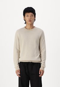 Beige textuurtrui met een ronde halslijn, geribbelde boorden en zoom. Gepaard met losse zwarte broeken. Effen achtergrond.