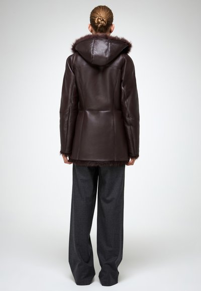 Giacca di pelle marrone con cappuccio e rifiniture in pelliccia, caratterizzata da un design aderente e maniche lunghe. Abbinata a pantaloni grigi a gamba larga.