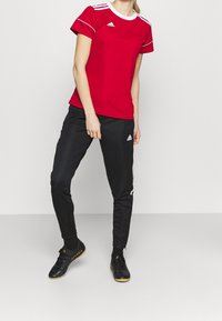 Červené sportovní tričko s bílými akcenty, kombinované s černými zúženými kalhotami s bílým logem. Černá obuv s texturovaným povrchem.