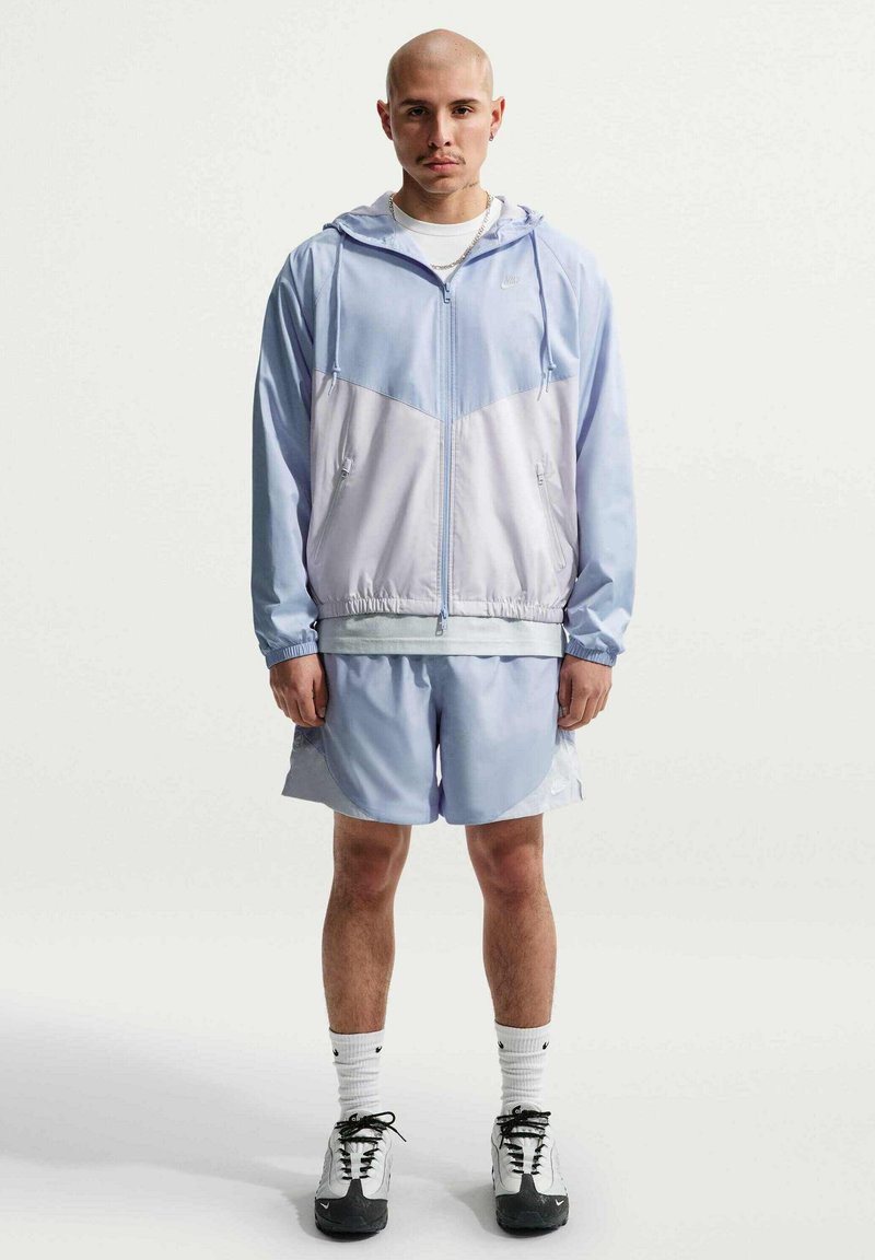 Adulte debout, les bras le long du corps, portant une veste zippée violet clair et blanc, un short assorti, des chaussettes blanches et des baskets noir et blanc.