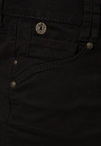 Mustat farkut, joissa on klassinen viisi-taskuinen muotoilu, messinkiset tarvikkeet ja hillityt ompeluyksityiskohdat taskuissa.