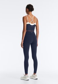 Nő hátulról állva, tengerészkék leggingsben, kék-fehér, keresztpántos crop topban és fehér sportcipőben, fehér háttér előtt.