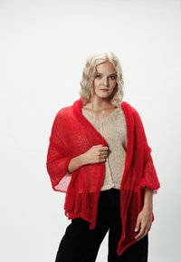 Czerwony szal z dzianiny o luźnym splocie z lekką teksturą, zarzucony na beżowy sweter, w połączeniu z ciemnymi spodniami. Modelka stoi na białym tle.