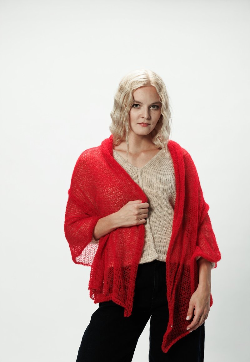 Czerwony szal z dzianiny o luźnym splocie z lekką teksturą, zarzucony na beżowy sweter, w połączeniu z ciemnymi spodniami. Modelka stoi na białym tle.