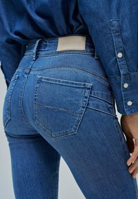 Temno modre jeans hlače z oprijetim krojem, poudarjenimi šivi in usnjenim logotipom na pasu, ter standardnimi zadnjimi žepi.