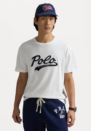 Camiseta de algodón blanca con el logo de "Polo Ralph Lauren" en azul marino, combinada con pantalones de chándal azul marino que presentan el texto "P.R.L." en blanco y cordones. Gorra oscura con logo.