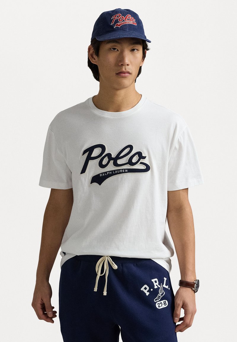 Balta kokvilnas t-krekla ar tumši zilu "Polo Ralph Lauren" logo, apvienota ar tumši zilu sporta biksēm ar baltu "P.R.L." tekstu un auklu. Tumša cepure ar logo.