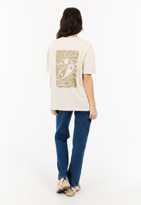 Crème-kleurig T-shirt met een grafisch ontwerp van een raket en krullen in beige. Gecombineerd met blauwe jeans en beige sneakers.