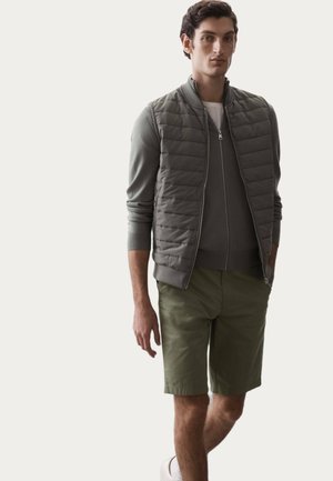 Leichtes, gepolstertes graues Westchen über einem eng anliegenden grünen Pullover, kombiniert mit olivgrünen Shorts. Weiße Sneakers runden den Look ab. Einfaches Design.