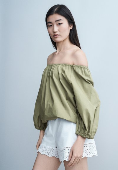 Jeune femme aux longs cheveux noirs portant un chemisier vert olive à épaules dénudées et un short blanc orné de dentelle ajourée, sur un fond uni.