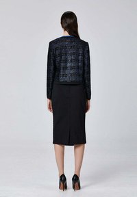 Jupe noire et veste texturée bleu marine à manches longues. Détail des chaussures montrant des escarpins noirs. Design simple et ajusté ; aucun motif visible.