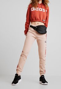 Person klädd i en röd avkortad Adidas-hoodie, beige Adidas joggingsbyxor med röda sidostråkar, svart midjeväska och svarta Adidas sneakers.