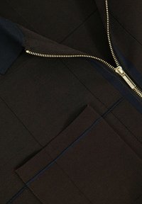 Närbild av ett mörkt plagg med en gulddragkedja och en ficka, som visar en texturerad textil med blå sömdekor.
