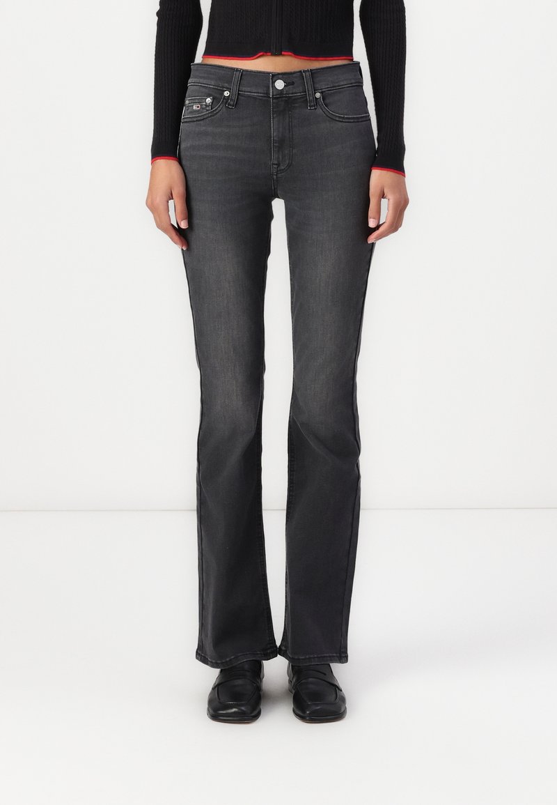 Personne portant un jean bootcut slim noir, des mocassins noirs et un pull court côtelé noir avec une bordure rouge, debout devant un fond blanc.