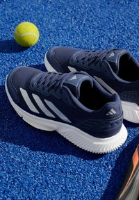 Два тъмносини и бели маратонки Adidas върху синя текстурирана спортна повърхност с жълта тенис топка наблизо.