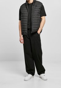 Černá vyp padací vesta s vysokým límcem, zipovým zapínáním a výrazným prošívaným vzorem, v kombinaci s černým tričkem a volnými černými kalhotami. Bílé tenisky.