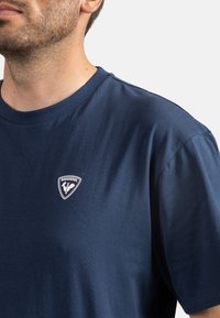 Námořnická modrá bavlněná tričko s kulatým výstřihem a krátkými rukávy, s bílou nášivkou loga Rossignol na levé části hrudi.