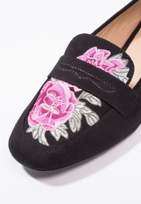 Mocassins en daim noir avec des roses brodées en rose et des feuilles vertes sur le dessus. Dotés d'une semelle plate et d'une bande décorative à l'avant.