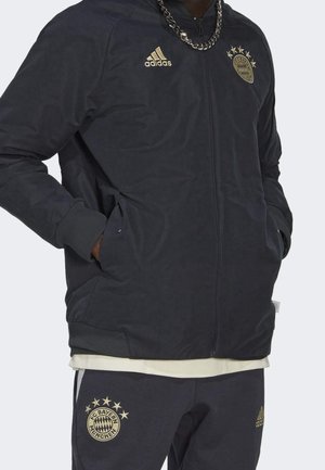 Personne portant une veste Adidas noire du FC Bayern Munich et un pantalon assorti avec des logos dorés et des étoiles, mains dans les poches, collier en chaîne argentée visible.