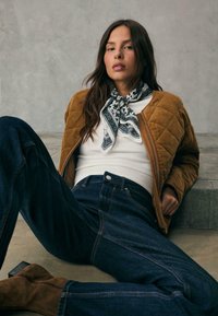 Brun quiltad bomberjacka över en vit skjorta, kombinerad med högmidjade mörkblå jeans. En svart-vit mönstrad scarf accentuerar outfiten.