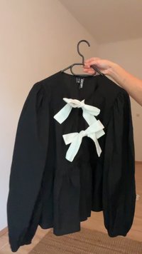 Blusa preta com mangas bufantes longas, adornada com dois laços brancos na parte da frente. Feita de um material leve. Projetada para uso casual.