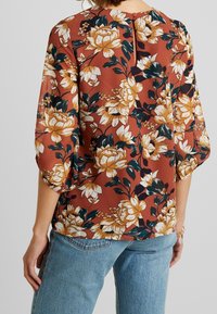 Blusa floreale in ruggine con fiori crema e verde scuro. Presenta maniche a sbuffo, una schiena a chiave e un tessuto leggero. Abbinata a denim.