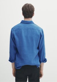 Man som står vänd bort med en blå långärmad skjorta med uppvikt ärm och svarta byxor mot en enkel vit bakgrund.