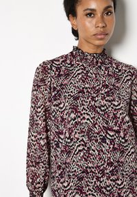 Blusa a maniche lunghe con collo alto arricciato, caratterizzata da un motivo astratto rosa e nero su uno sfondo chiaro. Realizzata in tessuto leggero.