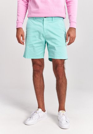 Miętowe szorty wykonane z lekkiego materiału, z płaskim przodem, bocznymi kieszeniami i podwiniętymi nogawkami, połączone z białymi trampkami.