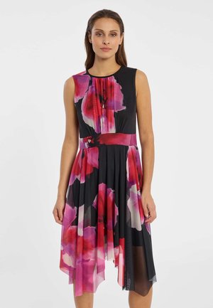 Frau trägt ein ärmelloses schwarzes Midikleid mit großem pink-rot-blumigem Muster und asymmetrischem Saum, steht vor einem schlichten Hintergrund.