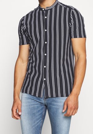 Hombre con camisa negra de manga corta con rayas blancas verticales y cuello mandarín combinada con jeans azul claro.