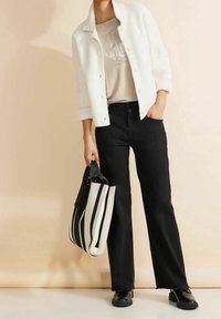 Weißer, flauschiger Button-up-Jacke über einem beigen Shirt, kombiniert mit schwarzen, weit geschnittenen Jeans und schwarzen Schuhen. Eine gestreifte Tote-Tasche wird in der Hand gehalten.