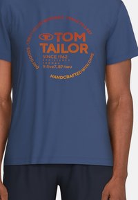 Camiseta de algodón azul con un estampado gráfico redondo en naranja que incluye detalles de la marca y cuidados, mangas cortas y una textura suave.