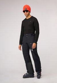 Zwarte lange mouwen shirt, zwarte sneeuwbroek met zilveren ritsen en textuuraccenten, feloranje beanie, zonnebril en skischoenen.