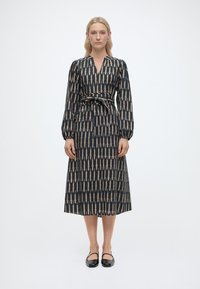 Γυναίκα που φοράει midi φόρεμα με μαύρο και μπεζ μοτίβο, ζώνη στη μέση και μακριά μανίκια, σε συνδυασμό με μαύρα παπούτσια Mary Jane.