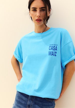 Donna con capelli scuri che indossa una maglietta azzurro chiaro con il testo "Fresh Tacos Casa Maíz" e jeans blu, in piedi su uno sfondo beige.