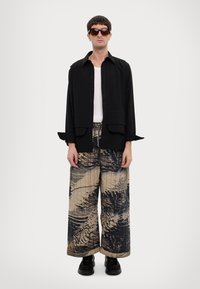 Black button-up shirt over a white tank, paired with wide, patterned pants in beige and dark tones, worn with black shoes and sunglasses.
