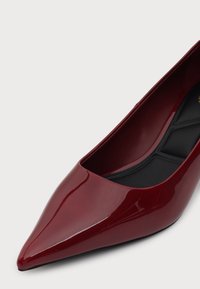 Chaussure à talon haut pointu rouge foncé brillant avec semelle intérieure noire matelassée sur fond gris clair.
