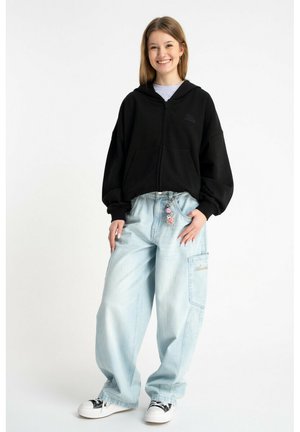 Młoda kobieta uśmiechająca się, ubrana w czarną bluzę z zamkiem, jasnoniebieskie, luźne jeansy z brelokiem oraz czarno-białe trampki, stojąca na białym tle.