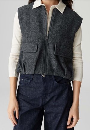 Gilet sans manches gris foncé zippé avec poches à rabat, porté sur une chemise crème à côtes à manches longues et un jean bleu foncé.