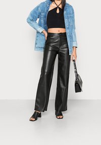 Pantalons larges en simili cuir noir, associés à un crop top noir et une veste en denim. La mannequin porte un petit sac à main noir et des sandales à talons.
