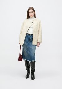 Vrouw die een crèmekleurig textuurjack draagt, een witte coltrui, een denimrok met rafels, zwarte laarzen tot de knie en een kleine donkerrode handtas vasthoudt.