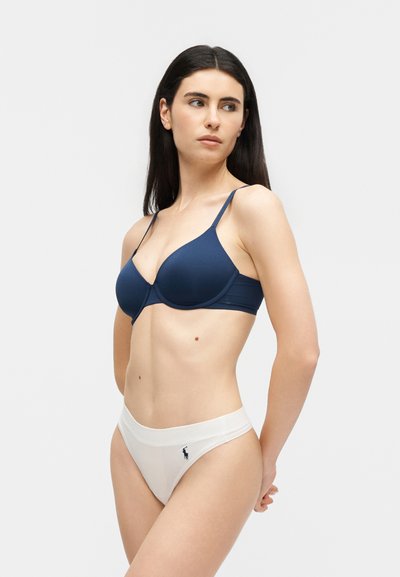 Femme portant un soutien-gorge bleu marine et un slip bikini blanc avec un petit logo noir sur la hanche gauche, debout devant un fond clair uni.