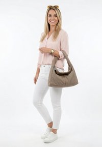 Glimlachende vrouw in een roze blouse en witte broek houdt een grote taupe schoudertas vast met een zonnebril op haar hoofd tegen een effen witte achtergrond.