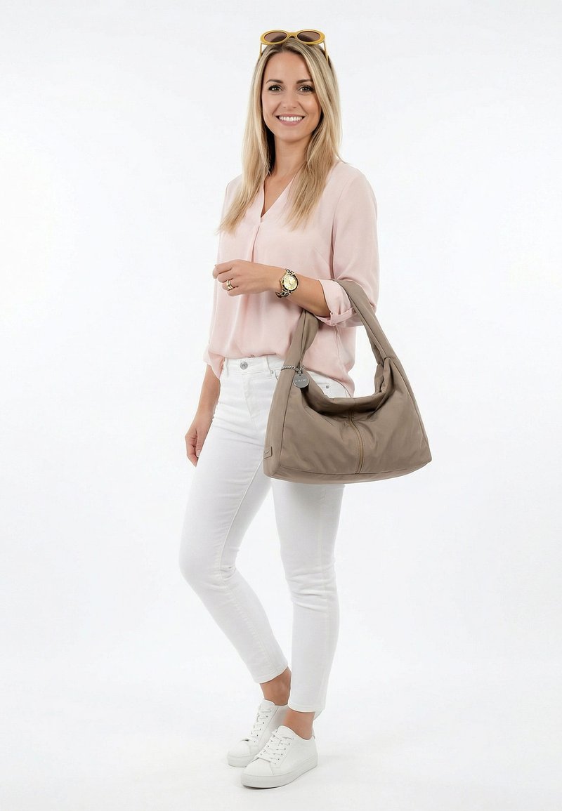 Glimlachende vrouw in een roze blouse en witte broek houdt een grote taupe schoudertas vast met een zonnebril op haar hoofd tegen een effen witte achtergrond.