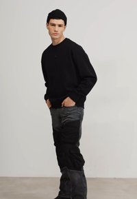 Felpa nera a girocollo abbinata a pantaloni cargo grigi e neri. Il modello è in piedi con le mani nelle tasche, indossando un berretto nero. Design semplice e moderno.