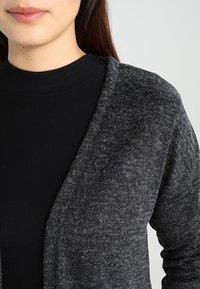 Szary dziergany sweter zapinany na guziki o miękkiej fakturze, z otwartym przodem i długimi rękawami, noszony na czarnym prążkowanym topie.