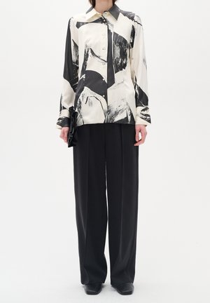 Persoon draagt zwarte broek met wijde pijpen en een blouse met lange mouwen en knopen met abstracte zwart-witte patronen.