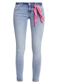 Vaqueros skinny de mezclilla azul claro con un diseño ajustado, con detalles desgastados y una bandana rosa con estampado atada a la cintura.